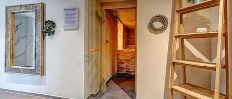 A hallway with a glass door leading to a room with a brick wall and a wooden door. There is a wooden shelf unit with a plant on top.