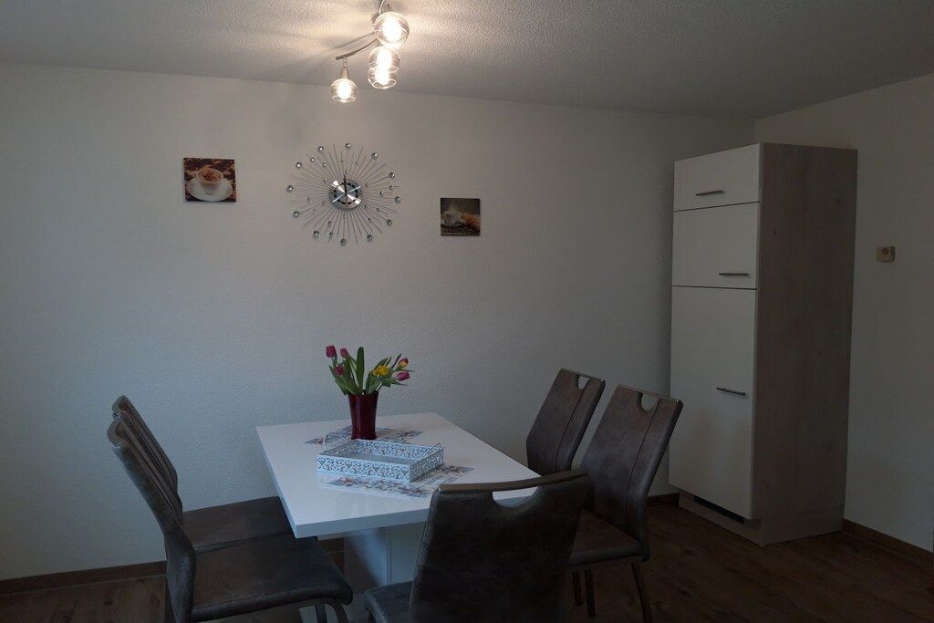 Un comedor con una mesa blanca, cuatro sillas, un florero con flores, un reloj y un armario.