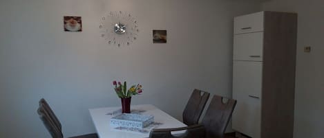 Un comedor con una mesa blanca, cuatro sillas, un florero con flores, un reloj y un armario.