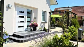 Een woonhuis met een witte gevel, een voordeur, een klein portaal met treden en een tuin met diverse planten en een groene parasol.