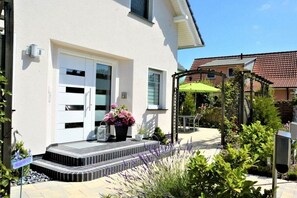 A residential building with a white exterior, a front door, a small porch with steps, and a garden area with various plants and a green umbrella. - Holiday apartment with 1 bedroom (Ostseeheilbad Zingst)