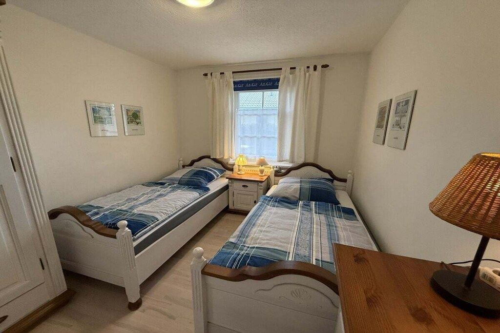 A bedroom with two single beds, a bedside table with a lamp, a window with curtains, and framed pictures on the wall.