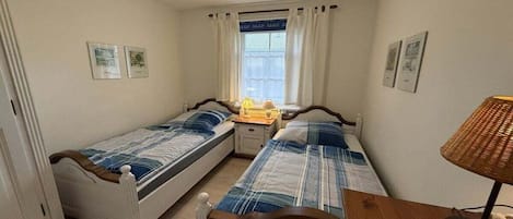 A bedroom with two single beds, a bedside table with a lamp, a window with curtains, and framed pictures on the wall.