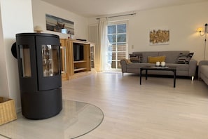 Ein modernes Wohnzimmer mit einem Glastisch, einer Couch mit Kissen, einem schwarzen Kamin und einem Fenster mit Vorhängen.