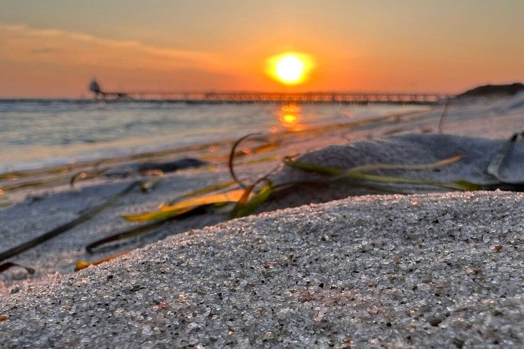 Eine Strandansicht mit Sonnenuntergang, Seetang am Sand und einem Steg im Hintergrund.