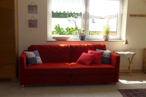 Ein rotes Sofa mit gemusterten Kissen, ein Fenster mit Pflanzen und ein kleiner Tisch mit einer Lampe.
