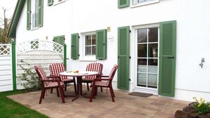Um pátio com mesa e cadeiras, um edifício branco com persianas verdes e um cercado branco.