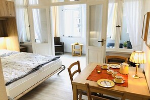 Ein Schlafzimmer mit Bett, Tisch mit Lampe und Stuhl. Es gibt ein Fenster mit Vorhängen, einen kleinen Tisch mit Pflanze und einen Stuhl in der Ecke.
