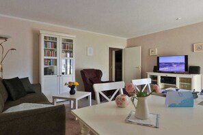 Un salon comprenant un canapé, une table blanche ornée d’un vase de fleurs, une bibliothèque, un téléviseur et une porte donnant sur une autre pièce.