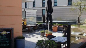 An outdoor seating area with tables, chairs, and umbrellas, overlooking a courtyard with a building in the background. - Holiday apartment with 2 bedrooms (Börgerende)