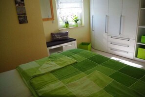 Une chambre à coucher avec un lit, une fenêtre ornée de plantes, une armoire et un banc.