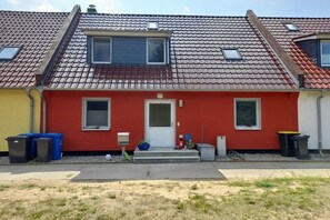 A red house with a tiled roof, a front door, and two windows. There are trash bins and a small garden area in front. - Holiday apartment with 3 bedrooms (Pruchten)