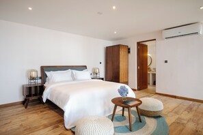 A bedroom with a bed, bedside tables, a wooden wardrobe, a round table with a vase, and a bathroom visible through an open door.