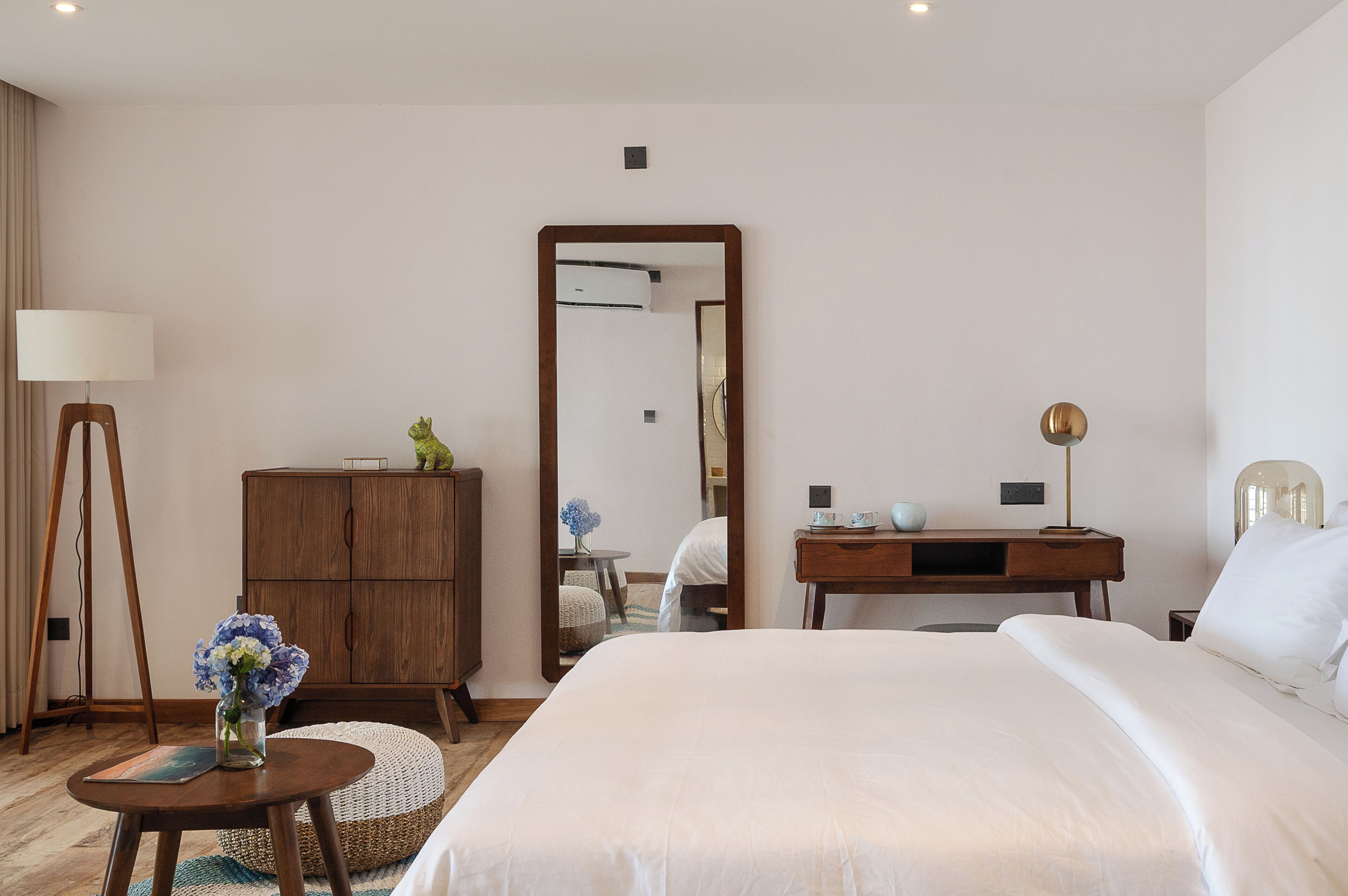 A hotel room with a bed, a wooden dresser, a desk, a lamp, and a mirror.
