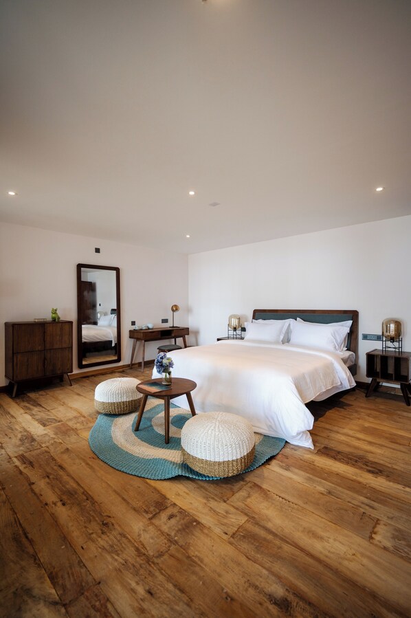A modern bedroom with a large bed, wooden flooring, a mirror, a desk, and a small seating area.