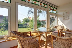 Ein Essbereich mit Korbstühlen und einem Glastisch, große Fenster mit Blick auf einen Garten und Bäume, sowie eine Wand mit einem dekorativen Element.