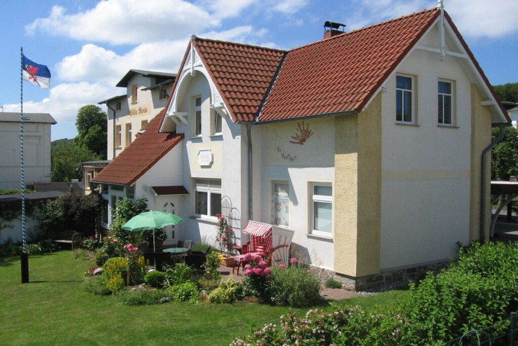 A two-story house with a red-tiled roof, a garden with flowers and a green umbrella, and a flagpole with a flag.