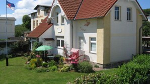 A two-story house with a red-tiled roof, a garden with flowers and a green umbrella, and a flagpole with a flag. - Holiday apartment Donnerkeil (Sassnitz)