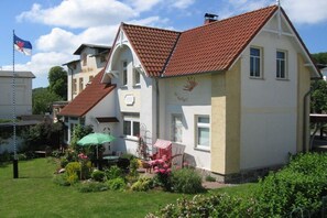 Een twee verdiepingen tellend huis met een rode pannendak, een tuin met bloemen en een groene parasol, en een vlaggenmast met een vlag.