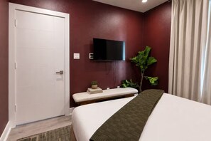 A hotel room with a bed, a bench, a TV mounted on the wall, and a door. - Moody Designer Retreat Near Greenville & Downtown (Dallas)