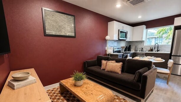 A modern living room with a dark sofa, a wooden coffee table, and a kitchen area in the background. - Moody Designer Retreat Near Greenville & Downtown (Dallas)