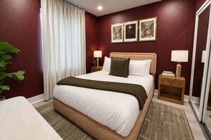 A hotel room with a large bed, two bedside tables with lamps, a window with curtains, and a plant. - Moody Designer Retreat Near Greenville & Downtown (Dallas)