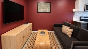 A modern living room with a flat-screen TV, a wooden coffee table, a sofa with cushions, a framed artwork on the wall, and a kitchen area with a microwave. - Moody Designer Retreat Near Greenville & Downtown (Dallas)