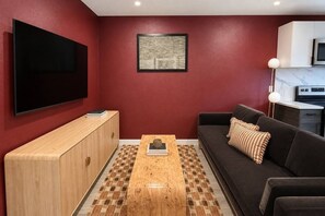 A modern living room with a flat-screen TV, a wooden coffee table, a sofa with cushions, a framed artwork on the wall, and a kitchen area with a microwave. - Moody Designer Retreat Near Greenville & Downtown (Dallas)