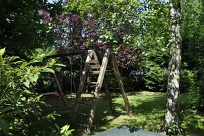 Un ensemble de balançoires en bois installé dans un jardin, avec une échelle, et entouré d’une végétation luxuriante et d’arbres.