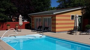 Una piscina con sillas de descanso y sombrillas, un pequeño edificio de fachada a rayas, y un cielo despejado.