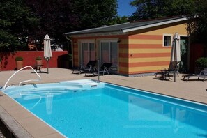 Una piscina con sillas de descanso y sombrillas, un pequeño edificio de fachada a rayas, y un cielo despejado.