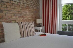 A hotel room with a brick wall, a bed with white bedding, striped pillows, a nightstand with a lamp, and a window with red and white curtains. - Holiday apartment with 2 bedrooms (Kühlungsborn)