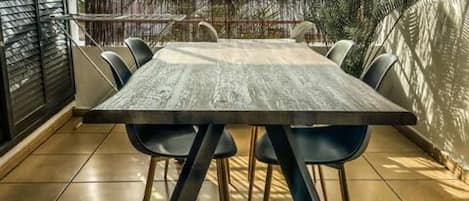 A rectangular dining table with four chairs, positioned on a tiled patio with a view of palm trees and a wooden fence.