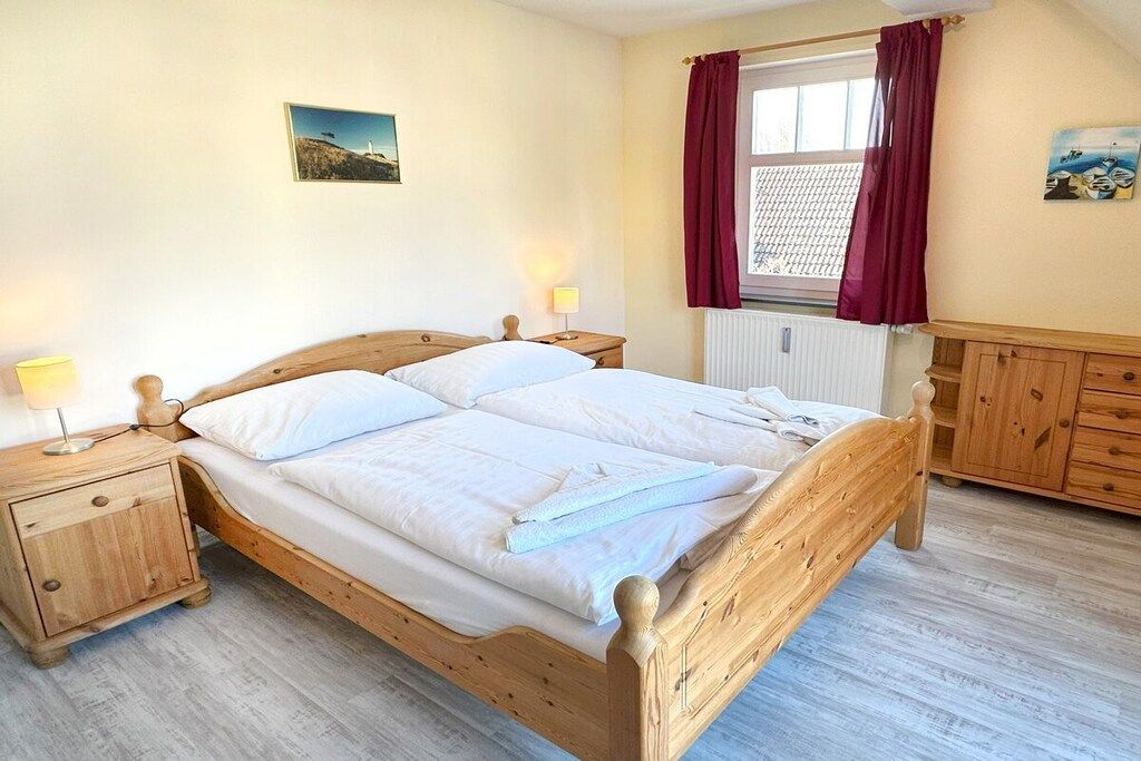 Ein hölzernes Bett mit weißen Leinen, ein Nachttisch mit Lampe, eine Holzkommode und ein Fenster mit Vorhängen.