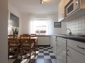 Eine Küche mit Schachbrettboden, Holzkufen, einem Esstisch, einem Fenster mit Vorhängen, einer Mikrowelle und einem Spülbecken.