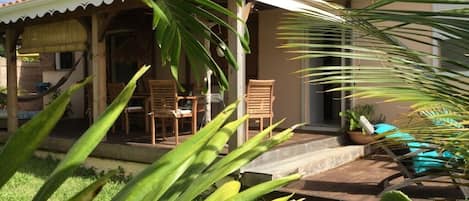 A covered outdoor seating area with chairs and a table, surrounded by lush greenery.