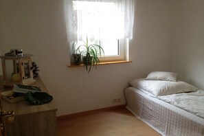 A bedroom with a single bed, a window with a plant, a bedside table with a lamp, and a small shelf with books and a decorative item. - Goodknecht Holiday Home (Rostock)