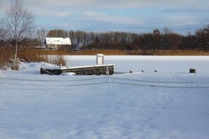 A snow-covered landscape with a dock extending into a frozen body of water, a small building with a snow-covered roof, and leafless trees in the background. - Holiday home in Pruchten with swimming pool (Pruchten)