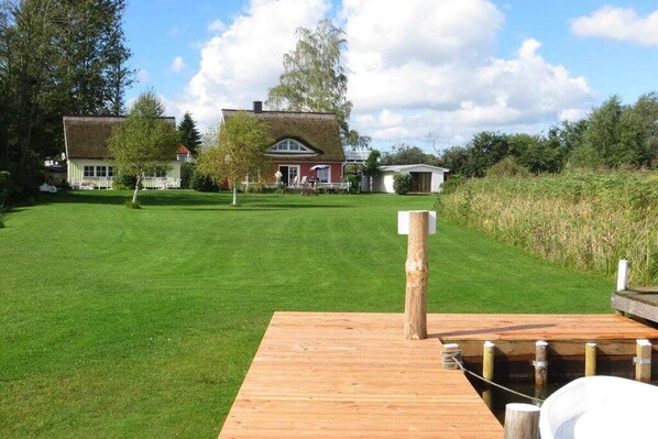 A grassy area with a wooden dock extending into a body of water, surrounded by trees and a house in the background. - Holiday home in Pruchten with swimming pool (Pruchten)