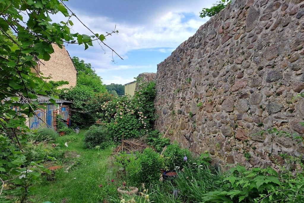 Un mur de pierre, un jardin avec différentes espèces de plantes et un bâtiment avec une porte bleue.
