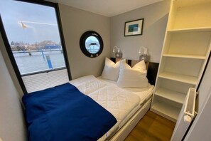 A bedroom with a bed, a window with a view of water, a circular mirror, and a white shelf unit.