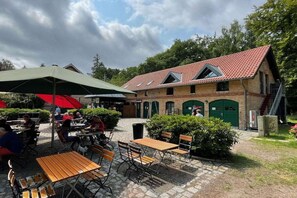 Een buitenruimte met tafels en stoelen, een overdekte parasol en een gebouw met een rode dakbedekking.