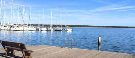 Un muelle de madera con un banco, cielo azul despejado y veleros amarrados en el agua.