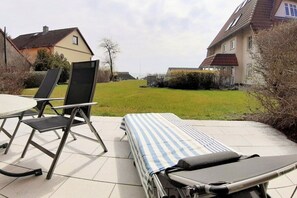 A patio with a lounge chair and a table, a grassy yard, and houses in the background. - Holiday apartment with 1 bedroom (Breege)