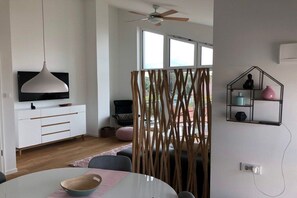 Ein modernes Wohnzimmer mit einem weißen Sofa, einem Esstisch mit einer Schüssel, einem Deckenventilator und einem Fernseher auf einer weißen Kommode.
