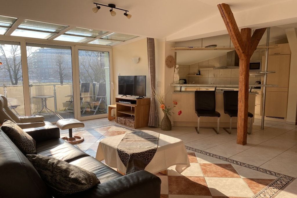 Ein modernes Wohnzimmer mit einer Couch, einem Couchtisch und einem Fernseher. Es gibt eine Essecke mit Stühlen und eine Küche im Hintergrund.