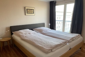 Un lit double avec une tête de lit sombre, une table de chevet, un tableau encadré au mur et une fenêtre avec des rideaux.