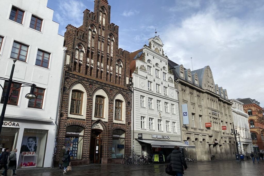 A historic building with arched windows and a clock tower, adjacent to modern shops and a white building with a sign.