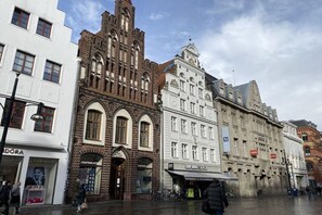 A historic building with arched windows and a clock tower, adjacent to modern shops and a white building with a sign. - Holiday apartment in Rostock with balcony (Rostock)