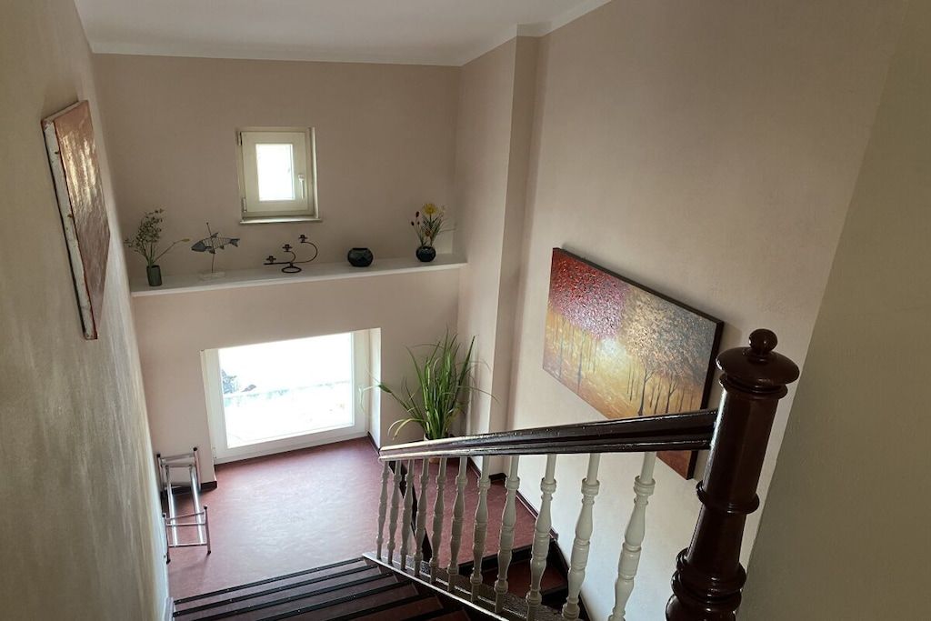 Un escalier doté d’une rampe en bois et de balustres blanches, menant à un palier avec une fenêtre et une étagère ornée d’objets décoratifs.